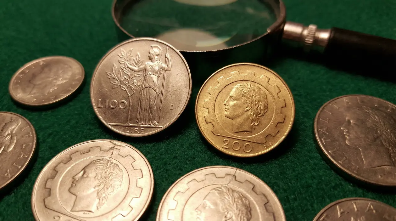 Monete in lire italiane con lente d’ingrandimento su panno verde, tra cui 100 e 200 lire da collezione