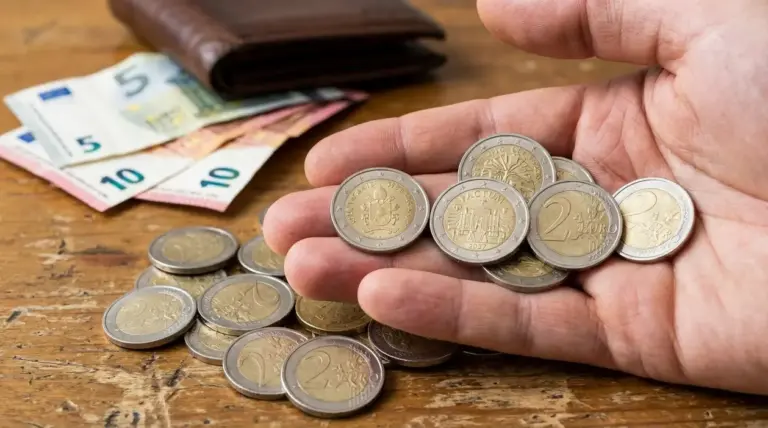 Mano con diverse monete da 2 euro su un tavolo, accanto a banconote e portafoglio