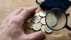 Mano che controlla alcune monete in euro con una lente d’ingrandimento su un tavolo di legno