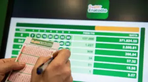 Mani che compilano una schedina del SuperEnalotto davanti a uno schermo con risultati e importi della vincita