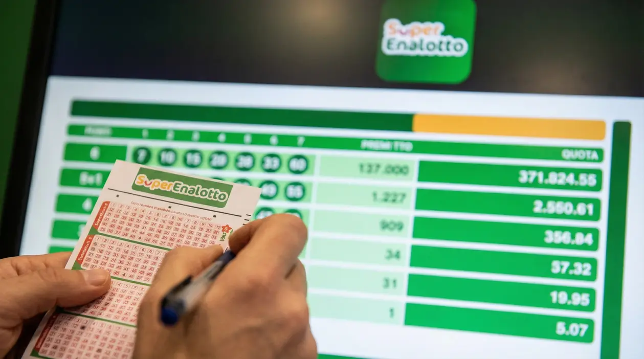 Mani che compilano una schedina del SuperEnalotto davanti a uno schermo con risultati e importi della vincita